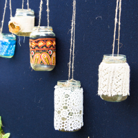 A display shows six glass jars hanging by twine against a dark blue background. Each jar is decorated with a different patterned fabric or lace. In the bottom left corner, a small patch of greenery is visible, along with a crochet piece draped partially in view, reminiscent of the 2013 Give and Get festival.