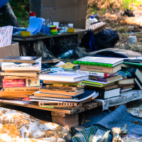 A stack of various books, notebooks, and folders sits on a makeshift pallet table outdoors at the Give and Get festival, surrounded by miscellaneous items including a cardboard box, pens, and a water bottle. The setting appears to be an informal outdoor workspace with grass and tarp visible in the background.