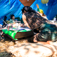 A group of people gathered under a blue tarp canopy at the 2013 Give and Get Festival, engaged in various activities. One person kneels at a low table covered with art supplies, while others sit on mats around the table. Sunlight filters through the trees, casting dappled shadows on the scene.