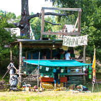 A wooden shack surrounded by trees with an upper deck, a light blue tarp for shade, and a sign reading "PANNAS" in large letters. People are gathered around and inside the shack, engaging in various activities at the 2013 Give and Get festival. The scene is bright and sunny, with grassy ground below.