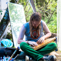 A person with long hair and a beard plays a stringed instrument while sitting on a blanket outdoors. They wear a sleeveless shirt with a circular design and green pants. Surrounding them are art supplies, a large mirror, and trees in the background. The scene evokes the relaxed, artistic vibe of the 2013 Give and Get festival.