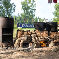 An outdoor makeshift kitchen at the 2013 Give and Get festival features a large metal stove and two big pots on a stone and brick hearth. A small fire burns underneath, with wooden shelter frames and drying laundry visible. In the background, a banner reading "KAFE3" is surrounded by trees and grassy landscape.