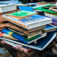 A large pile of books stacked haphazardly on a wooden surface. The books have various colorful covers and titles in different languages, including Latvian and English. Some spiral notebooks are also interspersed amongst the books from the 2013 Give and Get festival. The background is slightly blurred, showing more books and fabric.