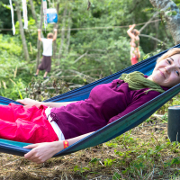 A woman with short hair smiles while lying in a blue hammock, wearing a purple shirt, red shorts, and a green scarf. Behind her at the 2013 Give and Get festival, a person balances on a slackline tied between trees, with another person nearby. The scene is set outdoors with lush green trees and a relaxed atmosphere.