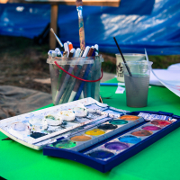 A table outdoors is covered with art supplies, reminiscent of the creative spirit at the Give and Get festival 2013. In the foreground, watercolor palettes burst with various colors. Nearby, a plastic cup holds water and paintbrushes, while a second cup contains murky water with a brush in it. Background includes a blue tarp and a whiteboard.