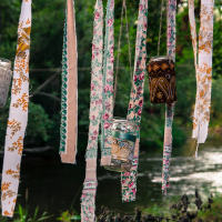 Glass jars and fabric strips hang from tree branches near a peaceful river, creating a whimsical and artistic atmosphere reminiscent of the 2013 festival. The jars are painted with colorful designs, some filled with small objects, while the fabric strips display various patterns and colors.