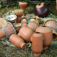 Various clay and wooden musical instruments, including drums, kalimbas, and jars, are laid out on a patterned cloth on grass. The instruments feature intricate designs and are used in traditional music. The setting appears rustic, with wooden planks and greenery visible in the background of the 2013 Give and Get festival.