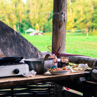 A rustic outdoor kitchen setup at the 2013 Give and Get festival features a gas stove, large mixing bowl, and several jars on a wooden counter. Various utensils and a coffee mug are scattered around. In the background, a lush green field with a volleyball net and trees is visible. Small lights hang from the wooden posts.