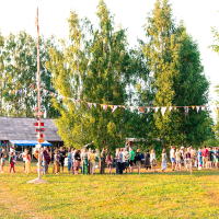A lively outdoor scene at the 2013 Give and Get festival in a rural area, featuring a crowd of people gathered under colorful bunting flags strung between tall trees. A rustic wooden building is visible in the background. The event takes place on a sunny day, creating a warm and festive atmosphere.