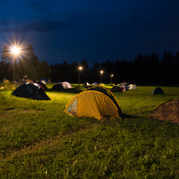A nighttime scene of a campsite during the 2013 Give and Get festival, with multiple tents pitched on a grassy field. Several tall lampposts illuminate the area with a warm glow, casting light and shadows on the tents and grass. The sky is dark blue, indicating it is well into the night, with trees silhouetted in the background.