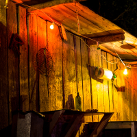A rustic wooden shack is dimly lit by colorful string lights, including red, yellow, and green bulbs. A striped shirt from the 2013 Give and Get Festival hangs on the left wall, and various items, including a wire rack and a plastic bottle, are attached to the walls. The scene is set against a dark, night-time background.