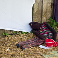 A homemade scarecrow, made from a fabric doll dressed in black, red, and striped clothing, leans against a weathered wooden wall. Surrounded by hay and green plants nearby, it evokes memories of the 2013 Give and Get festival. A white sheet is positioned in the background with a pair of red shoes near its feet.