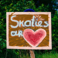 A handmade wooden sign is displayed against a backdrop of lush greenery at the 2013 Give and Get festival. The sign reads, "Skaties ar" in white, with a large red heart painted below the text. It has a rustic appearance, with visible brushstrokes and a brown background.