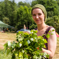 A woman with a calm expression stands outdoors holding a bunch of fresh green leaves. She wears a sleeveless black and red patterned top and has a green headscarf. Behind her, there are trees, a vehicle, and a green canopy tent, evoking the vibrant spirit of the Give and Get Festival 2013 in this forested, camp-like setting.