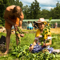 A man in orange shorts and a woman in a yellow top and blue skirt with a cowboy hat are crafting greenery wreaths outdoors. They are surrounded by lush trees and greenery at the Give and Get festival, with people standing near a large green tent in the background under a partly cloudy sky.