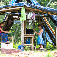 At the 2013 Give and Get festival, two women work under a makeshift wooden shelter with a tarp roof in a green outdoor setting. One woman sits weaving while the other stands, painting or staining cloth. Various clothes hang to dry above them, with trees and foliage visible in the background.