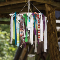 A homemade hanging mobile, reminiscent of the 2013 Give and Get festival, features numerous colorful fabric strips tied to a circular frame suspended by strings. A clear glass jar hangs below the frame, adding charm. The background reveals a rustic wooden structure with a green outdoors visible beyond.