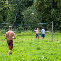 During the 2013 Give and Get festival, a group of shirtless people are playing volleyball on a grassy field surrounded by trees. The scene shows a net and several players actively engaged in the game, with two specifically focused on hitting the ball while others are positioned around them. Two individuals watch from the background.