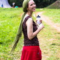 A woman smiles while standing in a grassy field with trees in the background. She wears a green headscarf, a sleeveless top with a black and red pattern, and a red skirt. She holds a water bottle in one hand at the 2013 Give and Get Festival. A dirt path and various outdoor equipment are visible behind her.