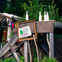 A rustic, outdoor cafe setup with a wooden stand adorned with painted white bottles and colorful bunting, reminiscent of the vibrant spirit of Give and Get festival. A handmade sign reads "Dad Cafe" with a green and red drawing, framed unevenly. Surrounded by lush greenery and a forested background, the weathered wooden structure exudes an informal charm.