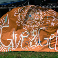 A large, orange banner with intricate, white and blue designs featuring swirls, stars, and an eye symbol at the top. The banner bears the painted words "Give&Get" in large, stylized white and red lettering. Displayed outdoors on a rustic wooden structure above green grass, it heralds the Give and Get festival 2013.