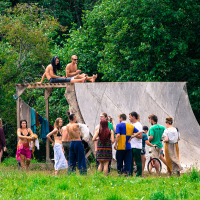 A group of people gather outdoors near a wooden ramp structure at the Give and Get festival in 2013. Some stand and chat, while others sit on top of the ramp. One individual holds a colorful sign with text on it. The scene is set in a lush, green environment with trees in the background and grass underfoot.