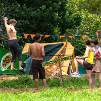 A group of four people at an outdoor campsite with orange and green flag bunting lines, reminiscent of the 2013 Give and Get festival. One person is balancing on a slackline while two others, shirtless, stand nearby. A woman in a yellow top is seated on a wooden post. A green and yellow tent and lush trees are in the background.