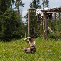 A person sits in a green grassy meadow taking photos with a camera. They wear a short-sleeved plaid shirt and sunglasses. Behind, there is a rustic wooden structure with a white cloth draped over it, reminiscent of the Give and Get festival. Tall trees are visible in the background under a partly cloudy sky.