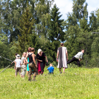 A group of people, including adults and children, engage in outdoor activities in a grassy area surrounded by dense trees during the 2013 Give and Get festival. Some hold sticks, suggesting a martial arts or reenactment practice. The weather is clear and sunny. One person leaps into the air, while others stand or watch the activity.