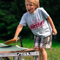 A young blond boy is standing on a wooden platform by a table tennis table at the 2013 Give and Get Festival. He is holding a table tennis paddle in one hand and a ball in the other. The boy is wearing a light blue T-shirt with red graphics and plaid shorts. The background shows greenery, suggesting an outdoor setting.