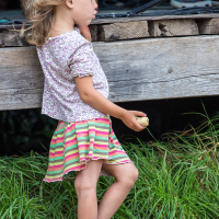 A young girl with light brown hair stands barefoot on grass, holding a small object. She wears a white shirt with small floral patterns and a colorful striped skirt in shades of pink and green. Her attention appears focused ahead, leaning slightly on a wooden platform at the 2013 Give and Get Festival.