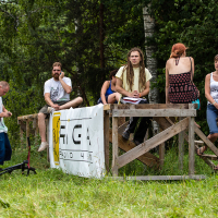 A group of people gather around a wooden platform in an outdoor setting surrounded by trees during the 2013 Give and Get Festival. Some are sitting on the platform, while others stand nearby. One person is writing in a notebook. There is a banner with text and a logo partially visible on the platform's side.