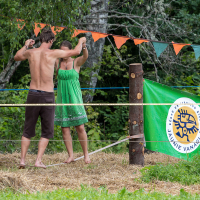 A shirtless man and a woman in a green dress are balancing on a slackline in a wooded outdoor area at the 2013 Give and Get festival. They hold hands for support. Orange and green triangular flags hang overhead, while a flag with a yellow and white emblem reading "Jaunie Vanagi" is displayed on a post to the right.
