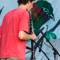 A man in a red T-shirt and khaki shorts plays a clarinet on stage at the 2013 Give and Get festival, facing away from the camera. Behind him is a microphone stand. Another person in a cap is blurred in the background. The backdrop features colorful illustrations of birds and greenery.