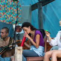 Three people sitting on stage appear to be preparing for a musical performance at the 2013 Give and Get festival. The man on the left, shirtless, holds a guitar. The woman in the center blows into a melodica, while the woman on the right, wearing a white embroidered top, looks ahead, holding a maraca. The background is colorful and eclectic.