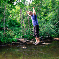 Amid the vibrant greenery of a dense forest during the 2013 Give and Get festival, a person balances on a wooden stick suspended by ropes over a shallow stream. Dressed in a blue shirt and black pants, they hold onto the ropes for stability while walking above the water. Lush foliage surrounds the scene.