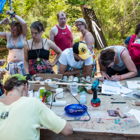 At the 2013 Give and Get festival, a group of people gather around a table outdoors under a blue tarp. Some are standing, while others are seated, focusing on various craft activities. The table is cluttered with art and craft materials like papers, tools, and rocks. Trees and greenery provide a scenic backdrop.