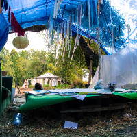 A vibrant campsite at the Give and Get festival is set under a blue tarp adorned with hanging ribbons and a lantern. In the foreground, a low table with papers and craft supplies sits on wooden pallets. A green couch is to the left. Tents and trees are visible in the background, creating a cozy, artistic outdoor environment.