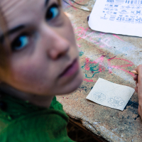 A woman with light-colored hair, wearing a green top, sits at a cluttered table looking up towards the camera. She holds a pen and is drawing intricate patterns on a small piece of paper. Nearby on the table is another paper with similar pattern sketches and various art supplies scattered around, reminiscent of the creative ambiance at the 2013 Give and Get festival.