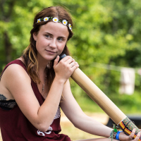 A woman with long brown hair, wearing a daisy flower crown, a maroon sleeveless top, and denim shorts, sits outdoors at the 2013 Give and Get Festival. She is holding a didgeridoo to her mouth, appearing focused. The background is lush and green with blurred foliage, suggesting a natural, wooded setting.