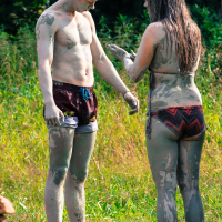 Two people are covered in mud, standing outdoors on grassy terrain at the 2013 Give and Get festival. The shirtless man on the left wears red and black shorts, while the woman on the right sports a chevron-patterned bikini. They appear to be talking or interacting, with trees and greenery visible in the background.