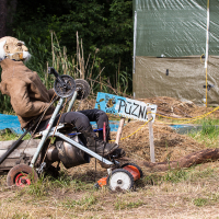 A scarecrow dressed in a brown jacket, black pants, and a worn mask with white hair sits on a makeshift vehicle made from scrap materials and an old lawnmower. Behind it, a hand-painted sign reads "PUZN," reminiscent of the 2013 Give and Get festival, surrounded by greenery and a tarp-covered structure. The ground is covered in dry grass.