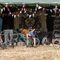 A bike repair stand is set up under a canopy at the Give and Get festival 2013. Various bicycles are being repaired or displayed, with tools and spare parts scattered around. Two individuals are working on bikes. Bunting and decorations hang from the canopy, while the ground is covered in dry grass.
