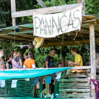 A group of people stands and sits under a wooden, open-sided shelter in a forested area during the 2013 Give and Get festival. A large hand-painted sign that reads "PANNCAKES" hangs at the front. Strings of lights and colorful fabrics decorate the space, giving it a festive look. Trees and greenery are visible in the background.