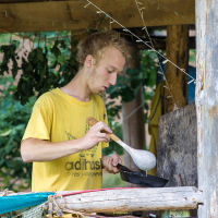 A young man with blond hair is under a rustic wooden shelter, wearing a yellow t-shirt with an illustration from the Give and Get Festival 2013. He is holding a large wooden spoon and appears to be cooking in a black frying pan. Various kitchen items and greenery surround the rustic setting.