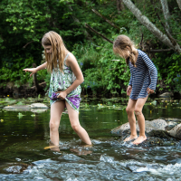 Two young girls are seen wading through a shallow, rocky stream at the Give and Get festival. The girl on the left wears a multicolored sleeveless dress, while the girl on the right sports a blue-and-white striped dress. Both are barefoot, carefully stepping on rocks amidst lush green trees and vegetation.