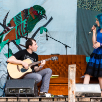 A man seated playing an acoustic guitar and a woman with blue hair standing with a microphone perform on an outdoor stage at the 2013 Give and Get festival. Behind them is a colorful bird mural and patterned fabric. An orange-brown couch and various musical equipment, including speakers, complete the scene.