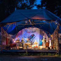 A band performs on an outdoor stage at night during the 2013 Give and Get festival. The stage, adorned with blue and white drapes and lights, hosts musicians playing drums, guitar, and keyboard. Trees frame the scene while an audience member watches from the right.