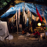 A cozy outdoor setup under a large blue tarp is visible, with various hanging white and red fabric decorations. A group of people are sitting on the ground around a lantern, engaging in activities reminiscent of the Give and Get festival. A white tent-like structure is on the left, and the surrounding area is dimly lit with a warm glow.