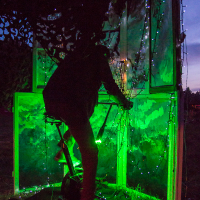 A silhouetted person operates an illuminated art installation at the 2013 Give and Get festival. The scene is lit with vibrant green lights, with string lights draped along the frame and surrounding structure. The dark background highlights the glowing elements of the installation beautifully.